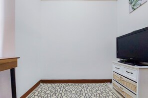 Quarto com televisão sobre um armário branco, tapete estampado e mesa de madeira.