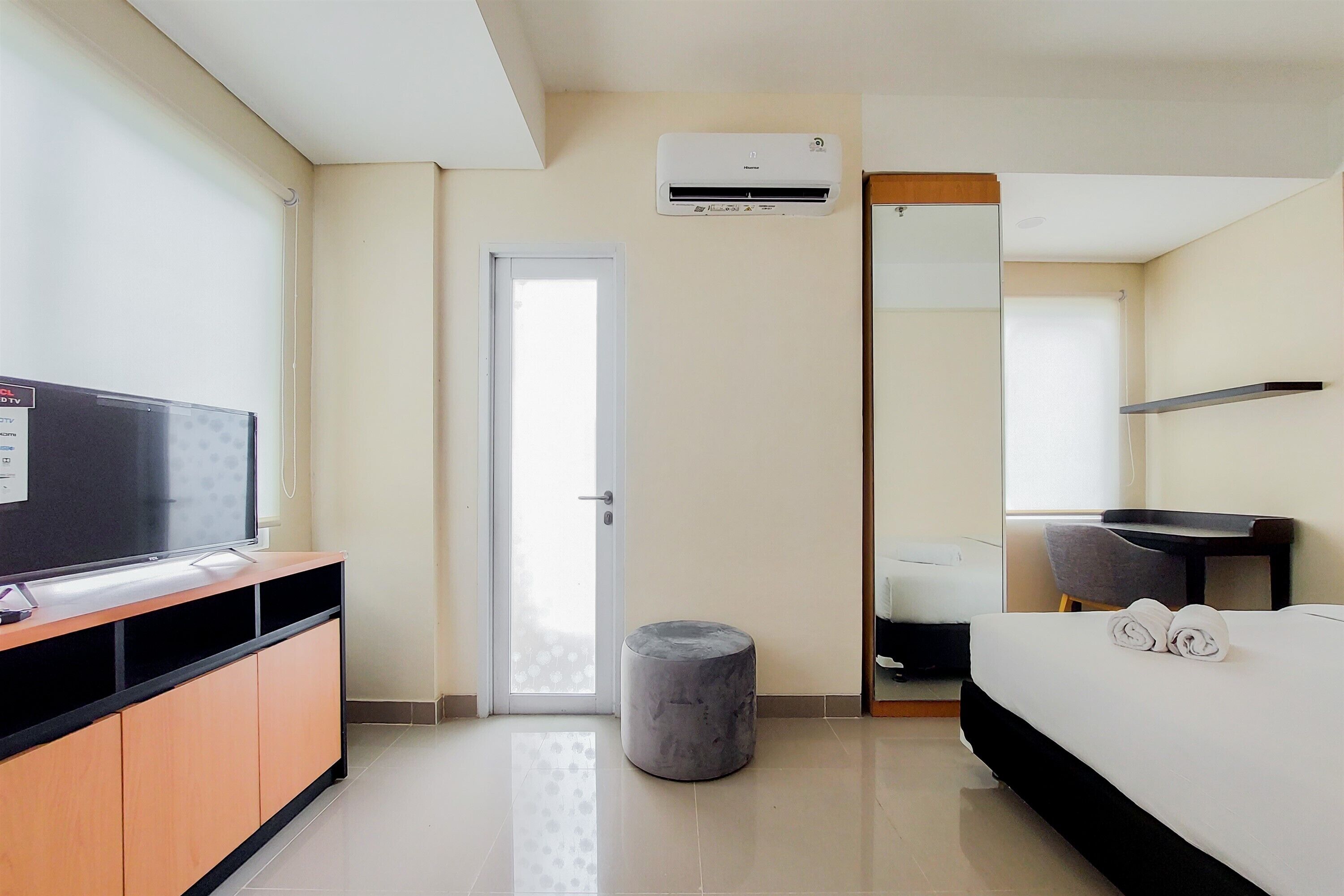 A hotel room with a bed, a TV on a wooden cabinet, a small round ottoman, an air conditioning unit, a door, and a mirror.