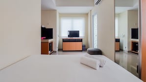 A hotel room with a bed, a television on a wooden cabinet, and a large window with blinds. - Homey And Comfort Studio Apartment At B Residence (Pagedangan)