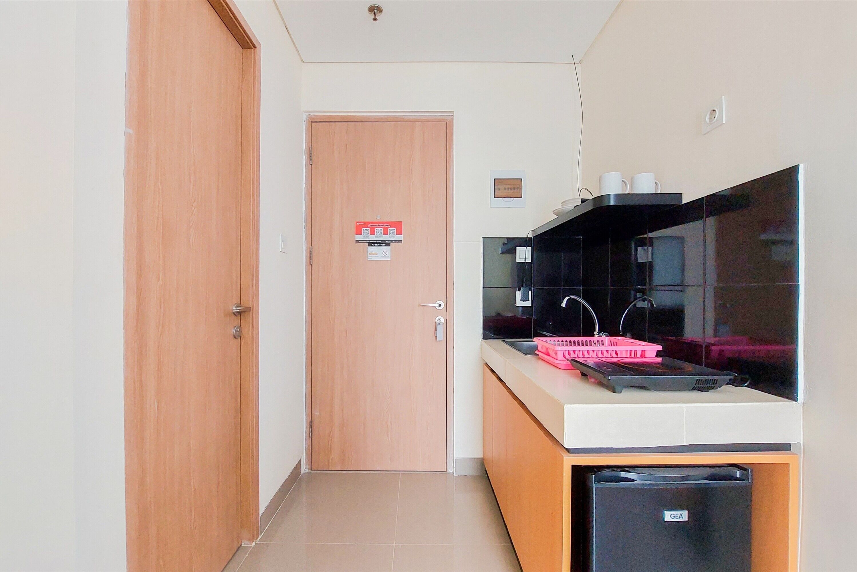 A compact kitchen with a microwave, sink, and a GEA refrigerator. A wooden door with a fire safety sign is visible in the background.