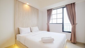 A modern hotel room with a large bed, a wooden headboard, and a window with curtains. - Minimalist 1Br With Extra Room At Evenciio Margonda Apartment (Depok)