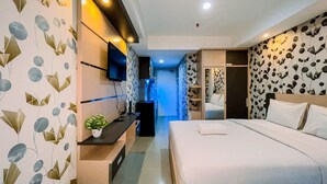 A modern hotel room with a bed, a TV mounted on the wall, a small plant on the nightstand, and a wardrobe with a mirror. - Simple And Cozy Studio Tamansari Skylounge Makassar Apartment (Makassar)
