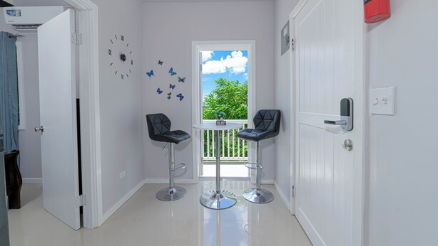A narrow hallway with two black bar stools and a small table. A white door is on the left, and a window with a view of greenery is on the right. - Modern, stylish, peaceful apartment with a great view! (Buckleys)