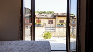 A bedroom with a large window, a bed, and a view of a courtyard with buildings and a fence.