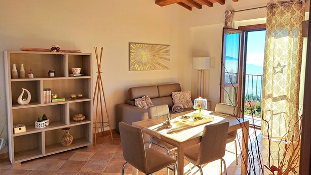 A cozy living room with a wooden ceiling, a sofa, a dining table with chairs, a bookshelf, and a view through a window.