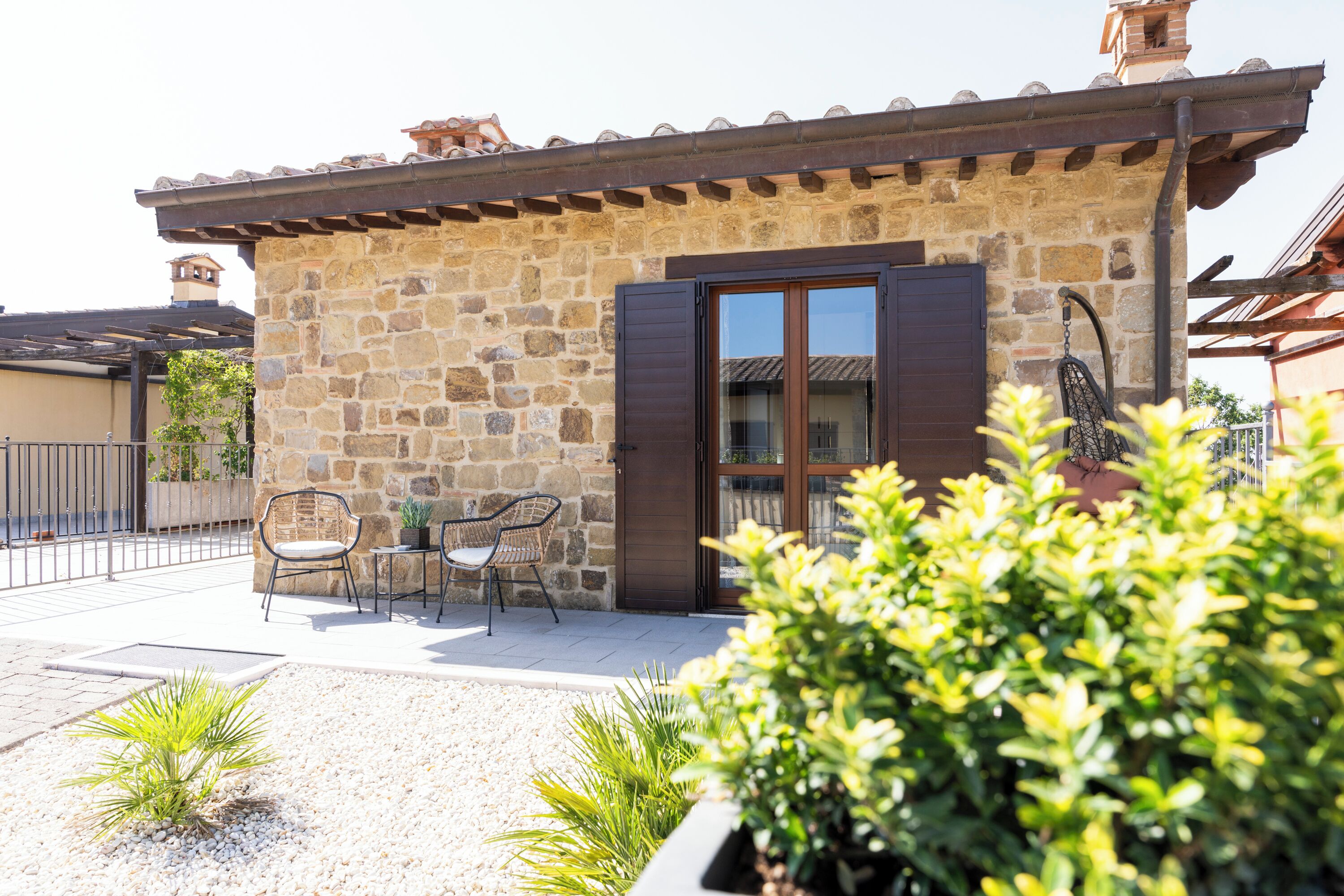 Un bâtiment en pierre avec un toit en tuiles, une porte et une terrasse agrémentée de chaises et de plantes.