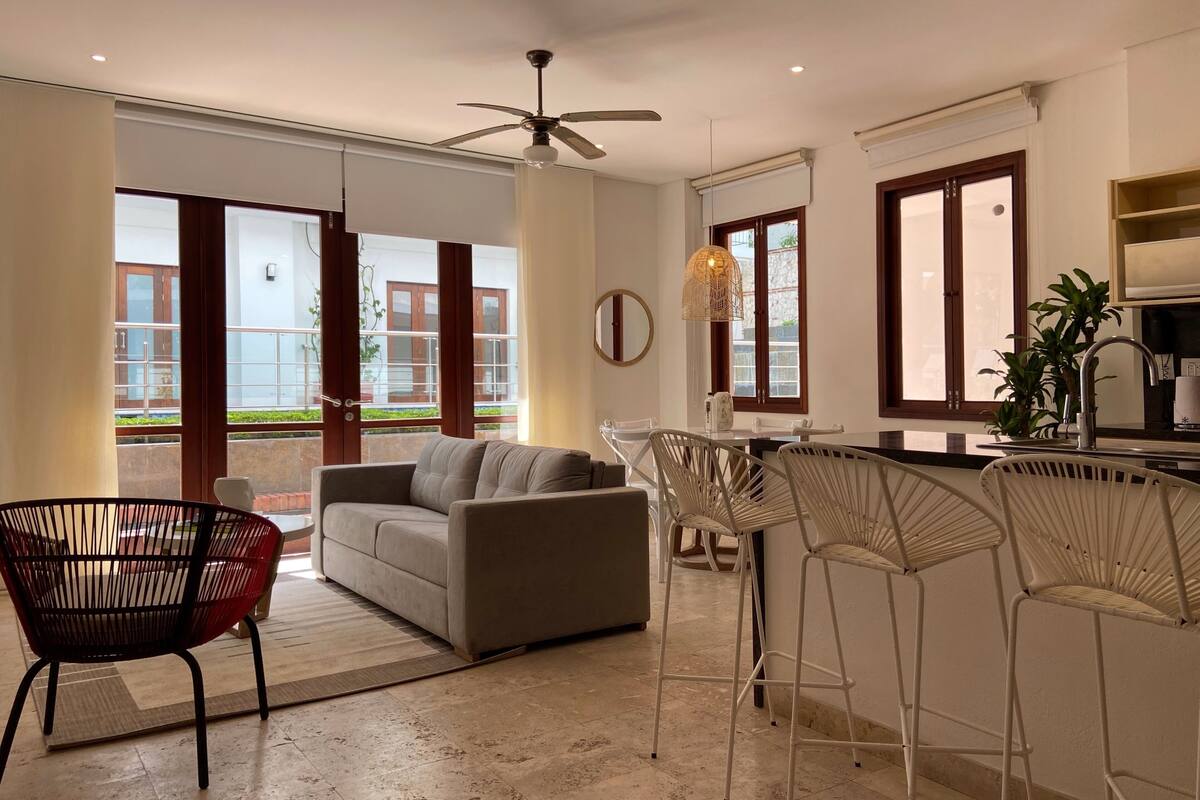A modern living room with a sofa, a dining table, and bar stools. There is a ceiling fan and a large window providing natural light.