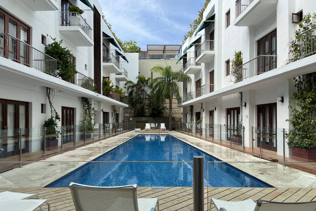 A swimming pool with lounge chairs, surrounded by white buildings with balconies and greenery.