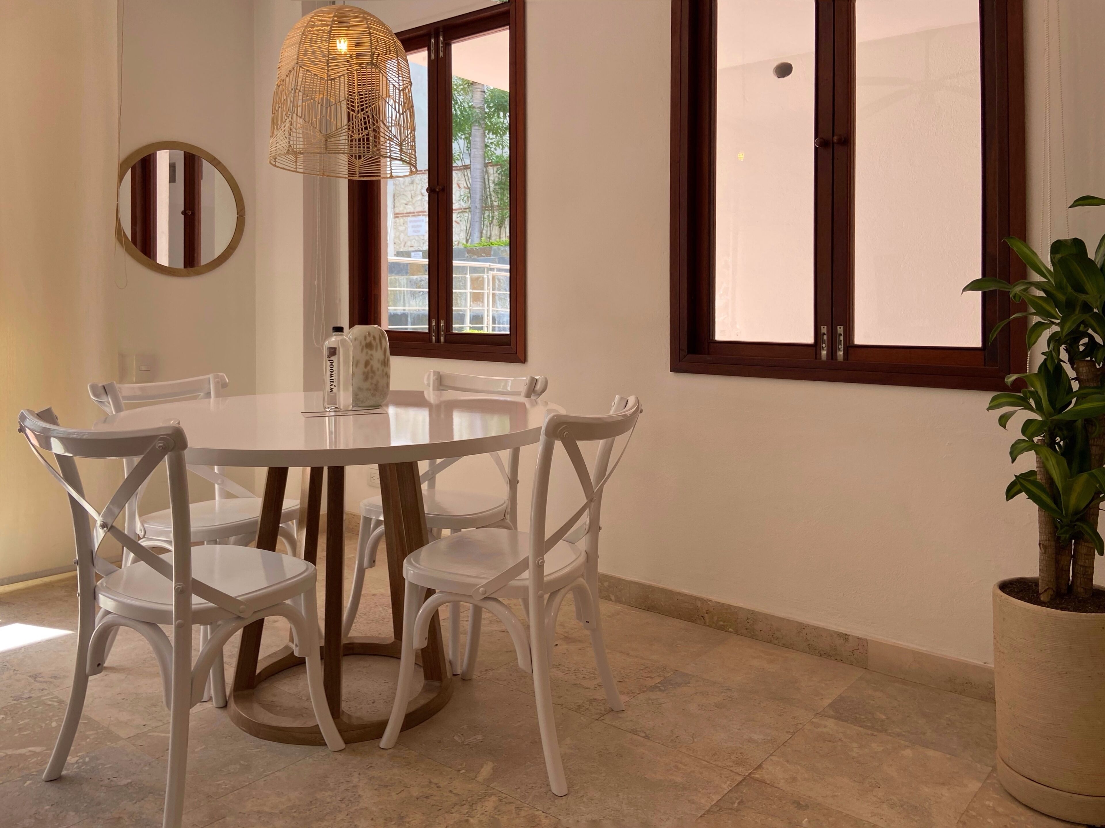 A dining area with a round table, white chairs, a hanging light fixture, and a potted plant.