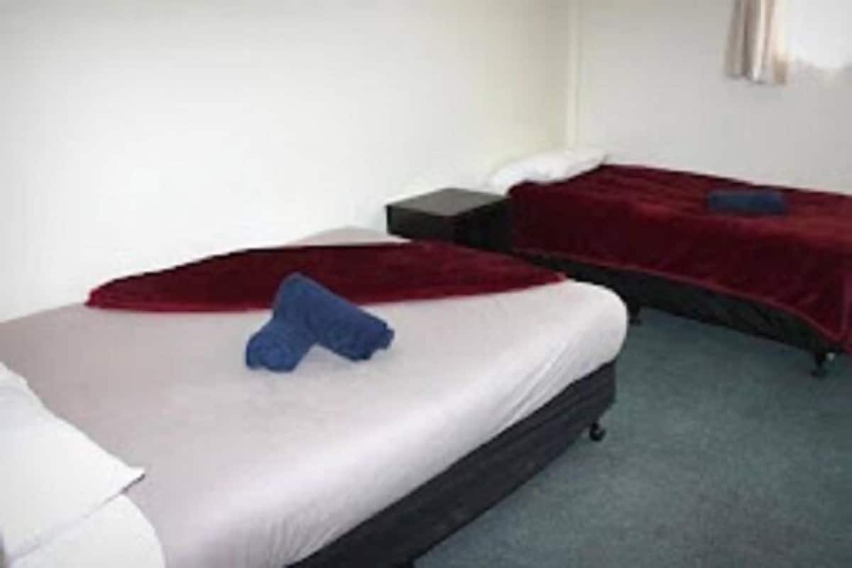 A hotel room with two beds, each with a red blanket and a pillow. There is a small bedside table between the beds.