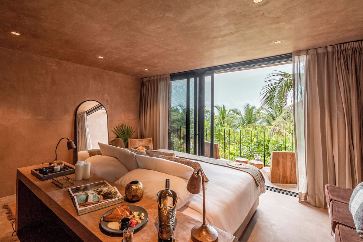 A modern hotel room with a large bed, a bedside table with amenities, a view of palm trees, and a balcony.