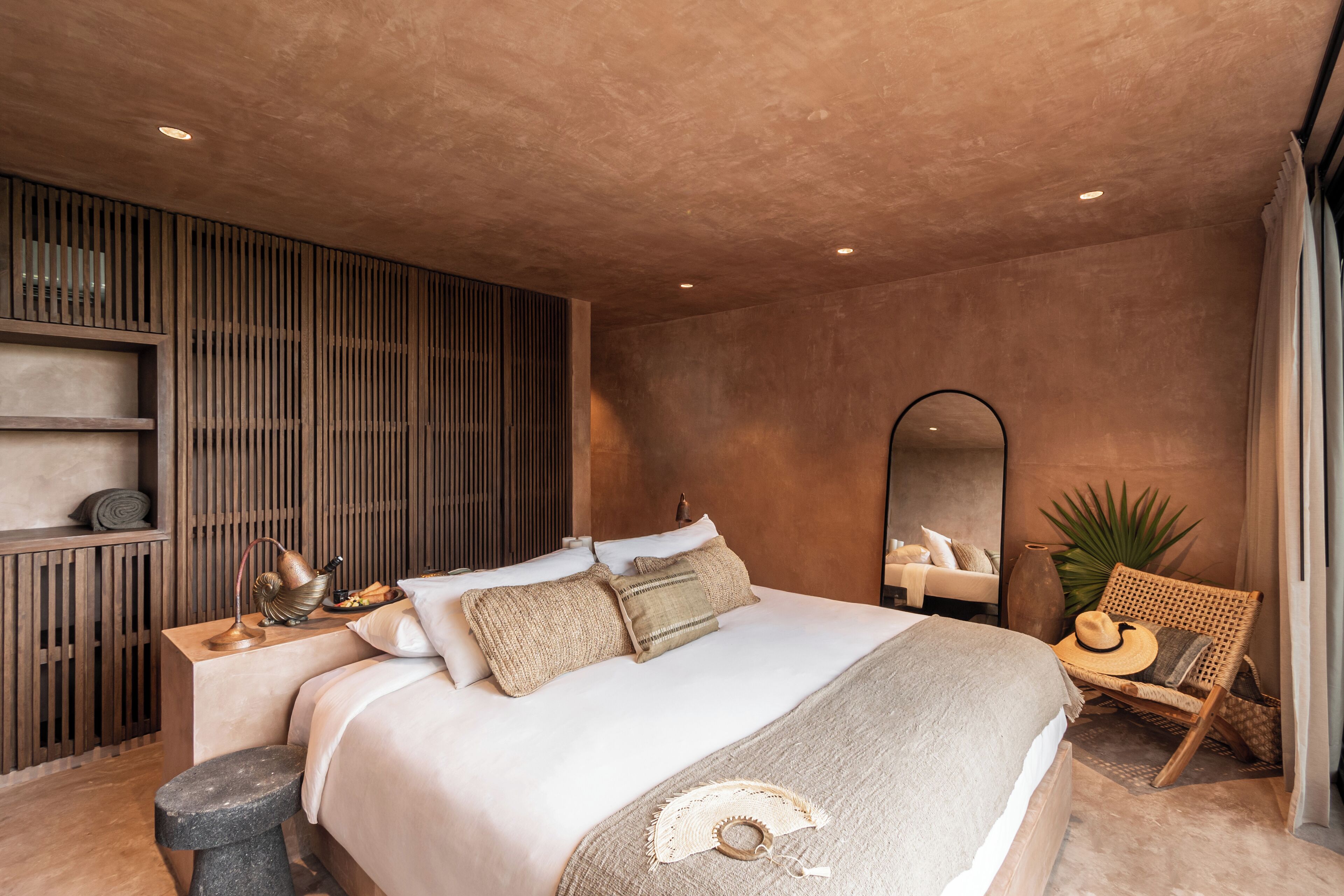 A modern bedroom with a large bed, a side table, a chair, a mirror, and a plant.
