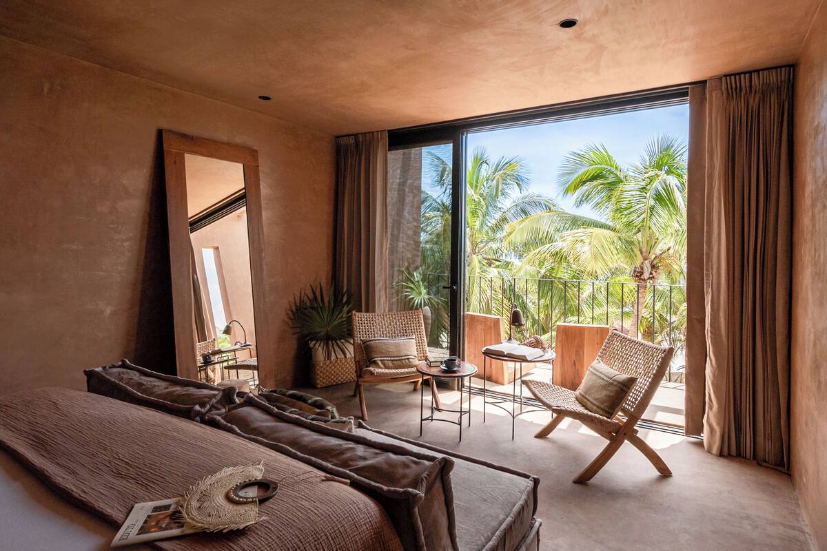A bedroom with a large bed, a sitting area with chairs, a small table, and a view of palm trees through a large window.
