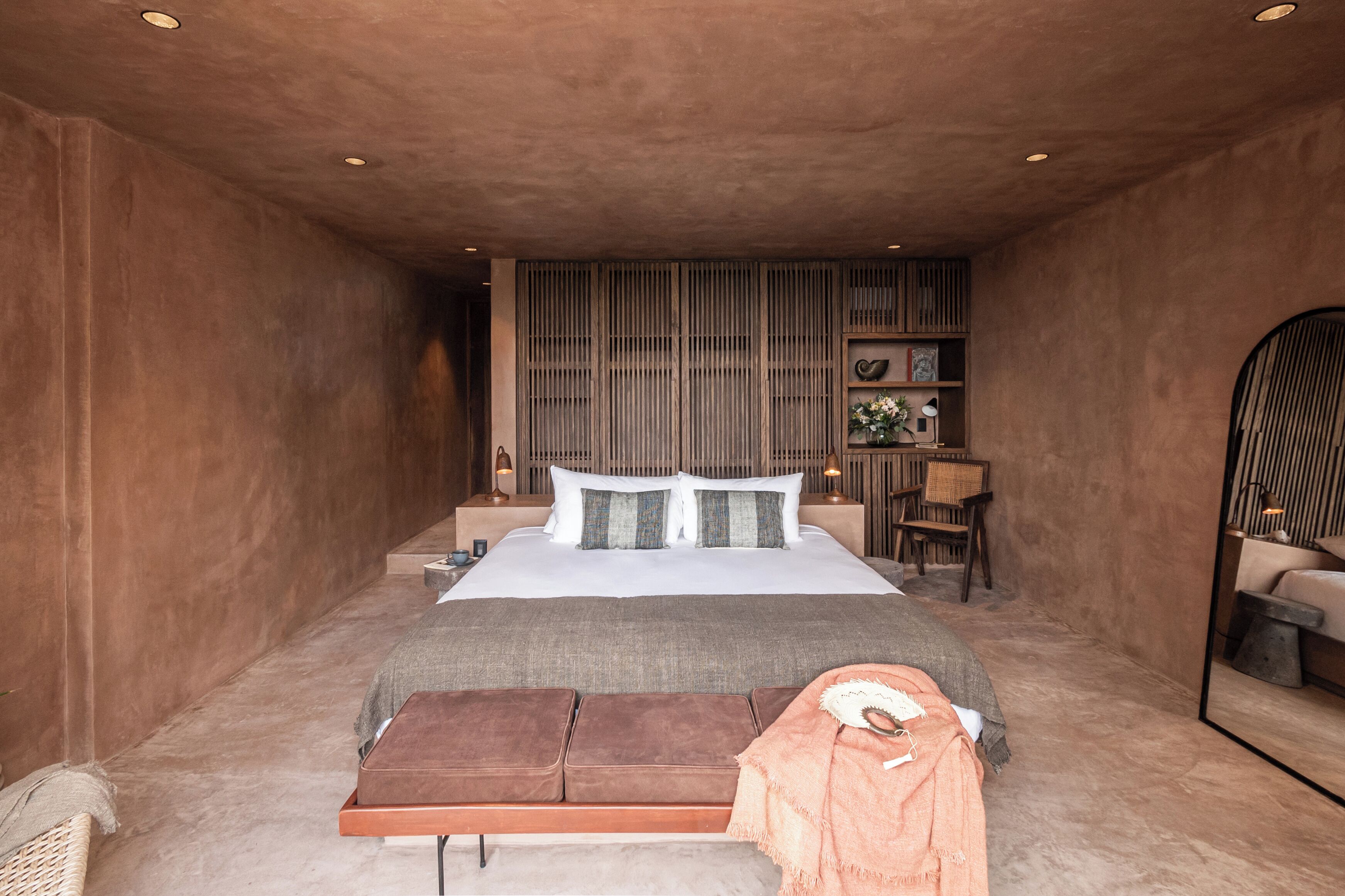 A bedroom with a large bed, a bench, a chair, and a small table with a lamp.