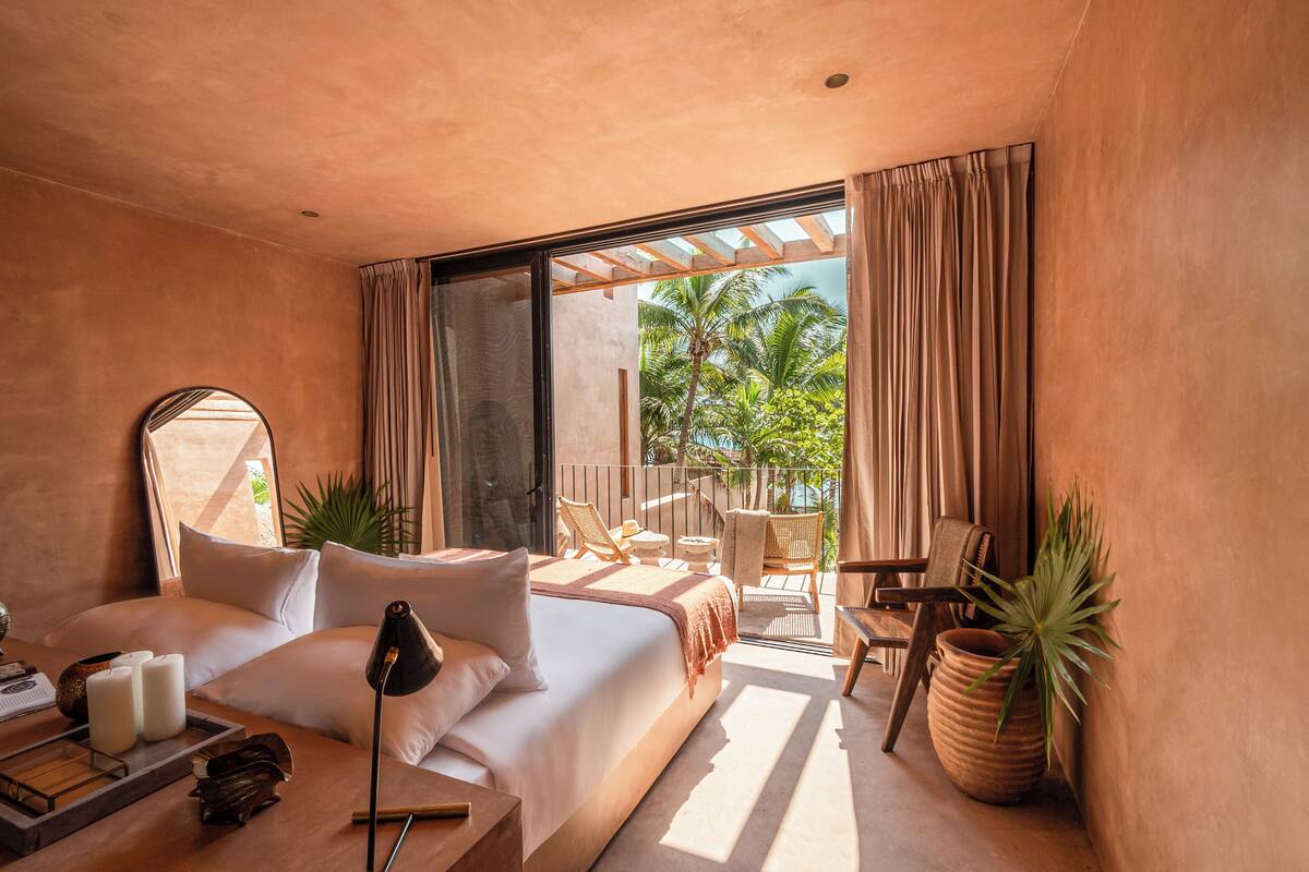 A bedroom with a large bed, a bedside table, a mirror, and a view of a balcony with outdoor furniture.