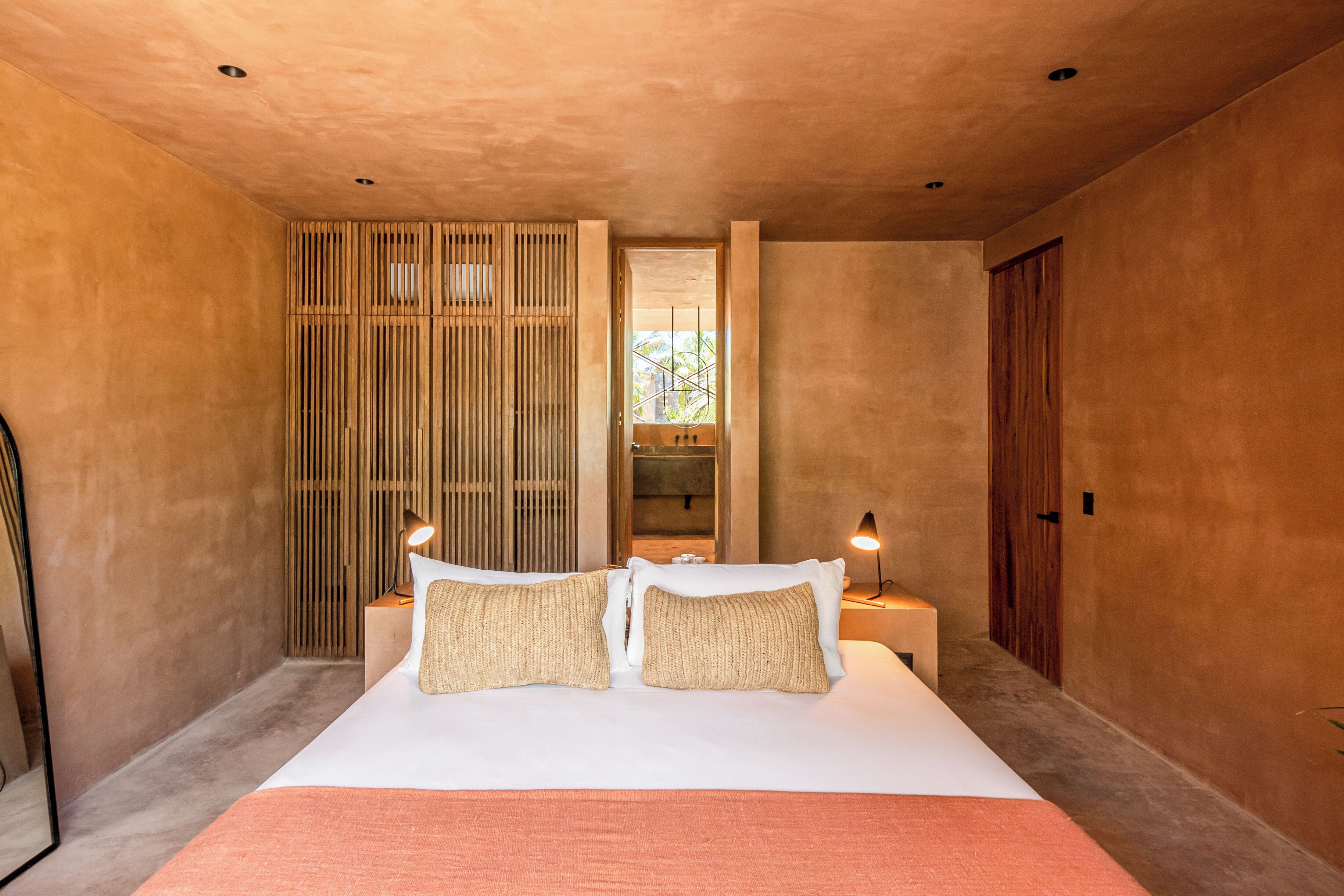 Quarto moderno com uma cama grande, divisória de ripas de madeira e um pequeno banheiro visível por uma porta.
