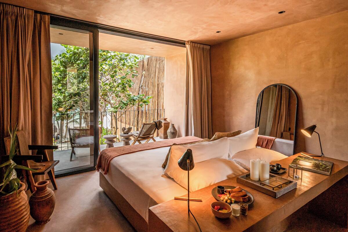 A modern hotel room with a large bed, a bedside table with a lamp, a mirror, and a view of the outdoors.