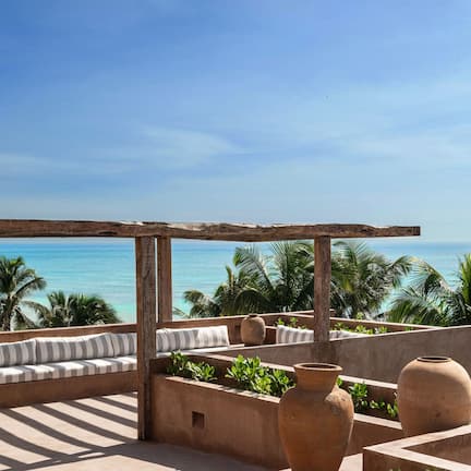 A terrace with a view of the ocean, featuring a wooden pergola, cushioned seating, and potted plants.