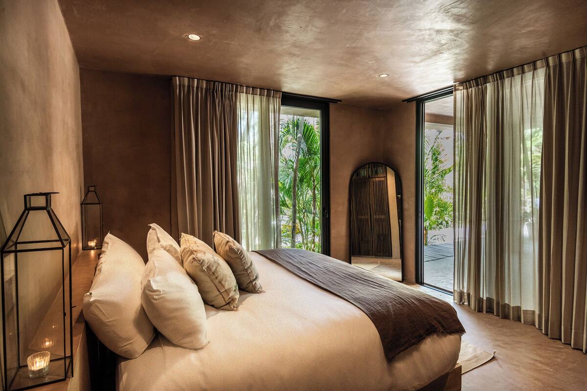 A modern bedroom with a large bed, multiple pillows, a bedside table with a candle, and a view of greenery outside.