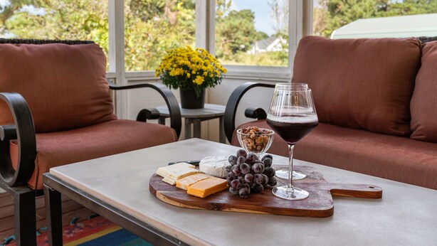 A table with a cheese board, grapes, and a wine glass in a room with brown sofas and a window with a view of trees and a house. - Meadow Farm in Holly Springs, NC (Holly Springs)