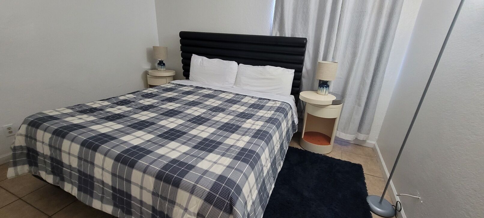 Ein ordentlich bezogenes Bett mit karierter Tagesdecke, zwei Nachttischlampen und einem dunklen Teppich auf Fliesenboden.