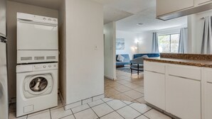 A compact kitchen with a built-in washer and dryer, a countertop, and a view into a living area with a sofa and a window.
