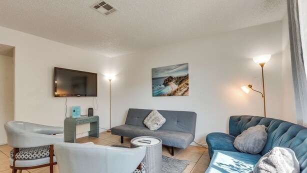 A modern living room with a grey sofa, a blue sofa, and a white armchair. There is a flat-screen TV mounted on the wall, a small table with a lamp, and a painting of a coastal scene.