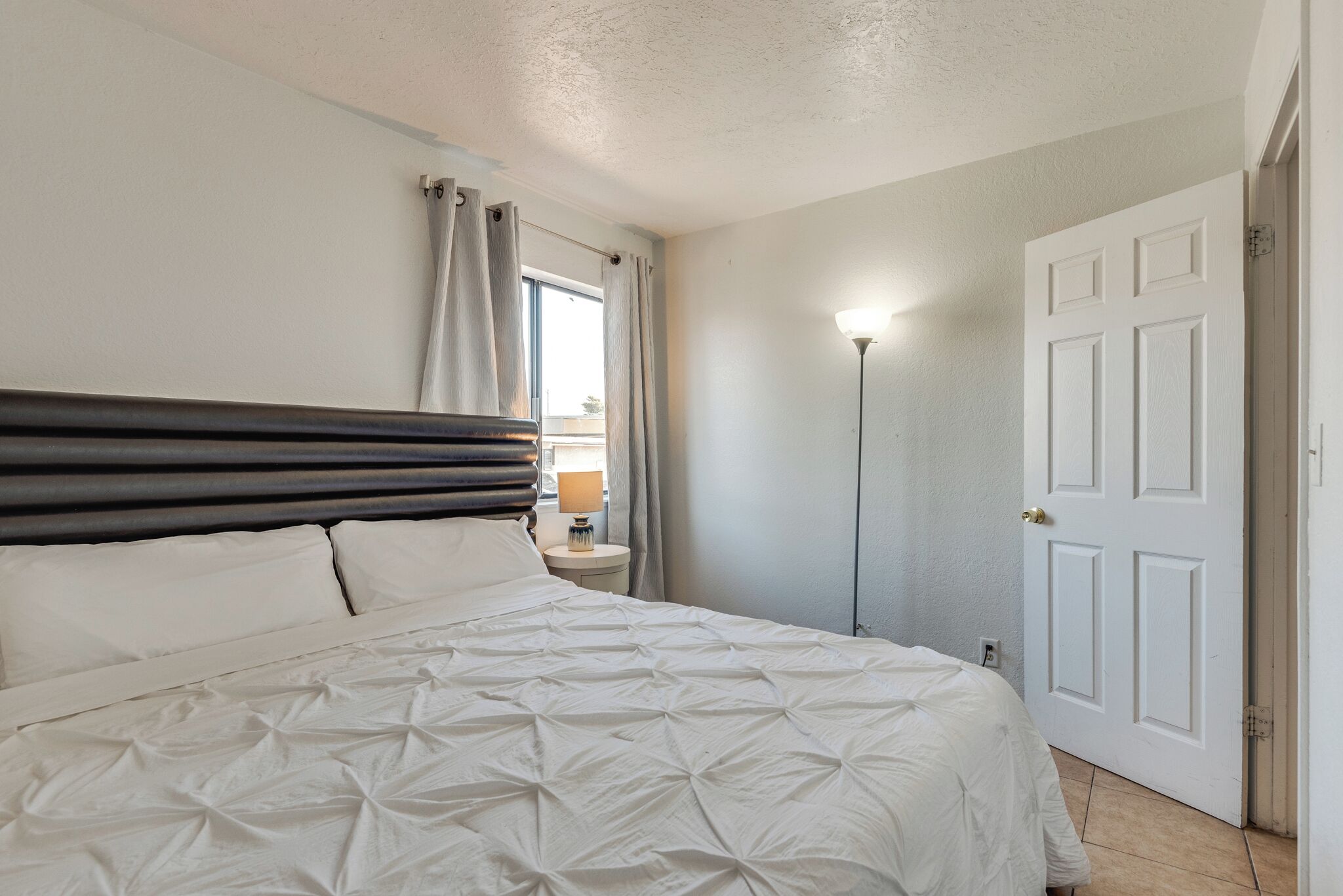 Ein Schlafzimmer mit Bett, Vorhängen, Nachttisch, Lampe und einer Tür.