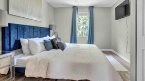 Une chambre à coucher avec un lit, une table de chevet, une lampe, un téléviseur et une fenêtre avec des rideaux.