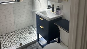 A modern bathroom with a white tiled shower, a white sink with a dark countertop, and a large mirror above the sink.