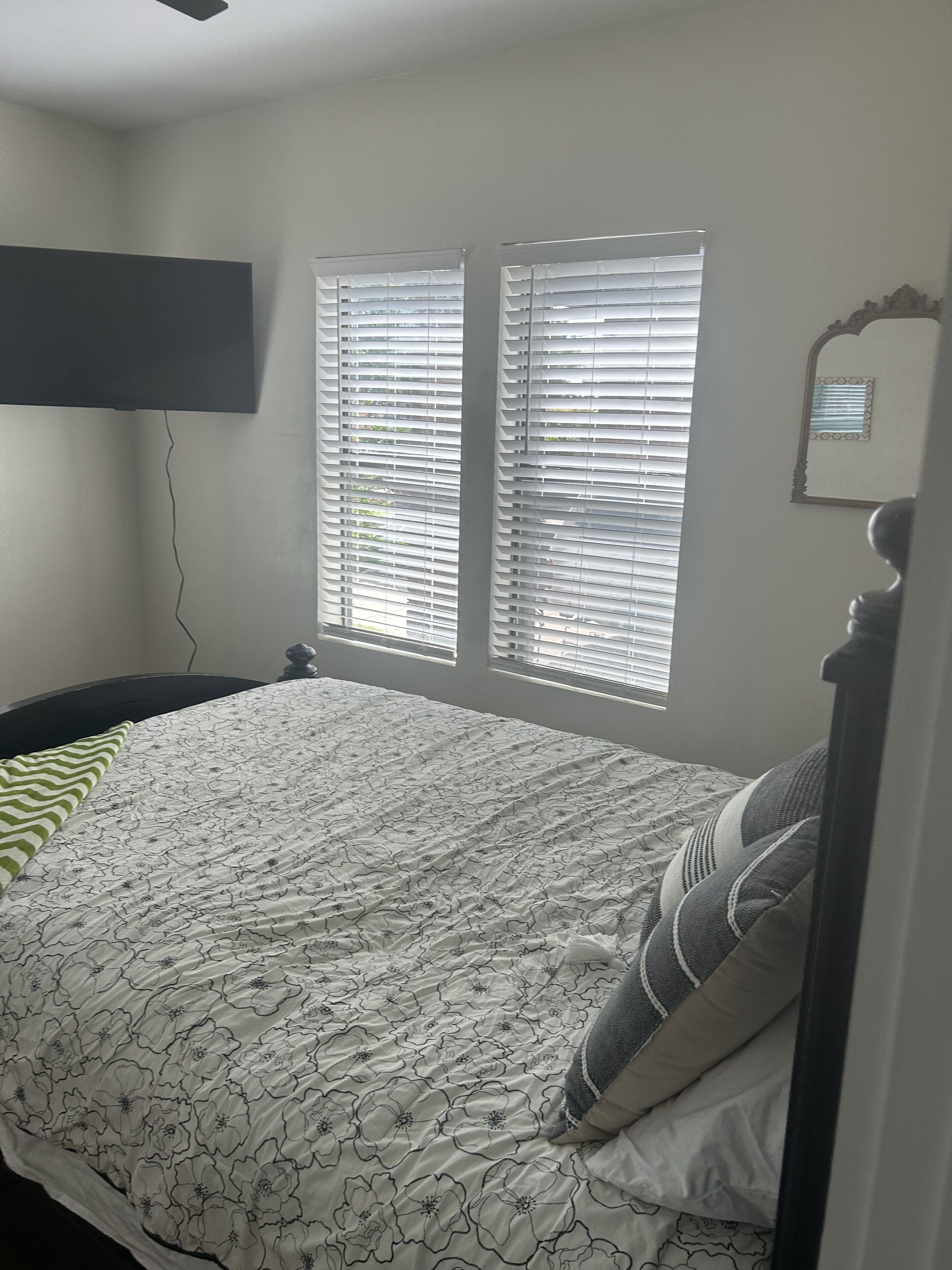 A bedroom with a bed, two windows with blinds, a wall-mounted TV, and a mirror.