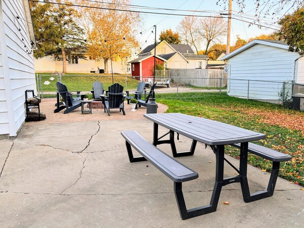 An outdoor patio with picnic tables and chairs, a small shed, and a fenced yard with trees.