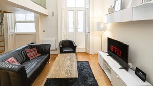 Un salon moderne avec un canapé en cuir noir, une table basse en bois, un fauteuil noir, un meuble TV blanc orné du logo Netflix et une grande fenêtre donnant sur l’extérieur.