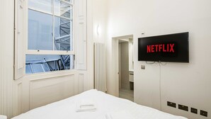 Une chambre d’hôtel avec un lit, une fenêtre, un radiateur, une porte et un téléviseur fixé au mur qui affiche « NETFLIX ».