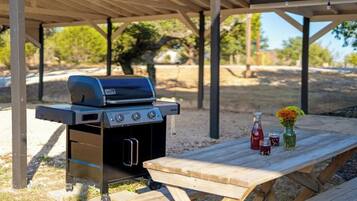 Una parrilla para barbacoa bajo un porche cubierto, con una mesa de picnic, un jarrón con flores y dos botellas.