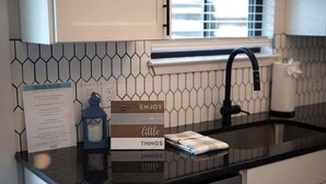 A kitchen with a black countertop, a white tiled backsplash, and a black faucet. There is a sign titled "HOUSE RULES" and a book titled "ENJOY THE little THINGS." - Fred's Joint With Hot Tub (Fredericksburg)