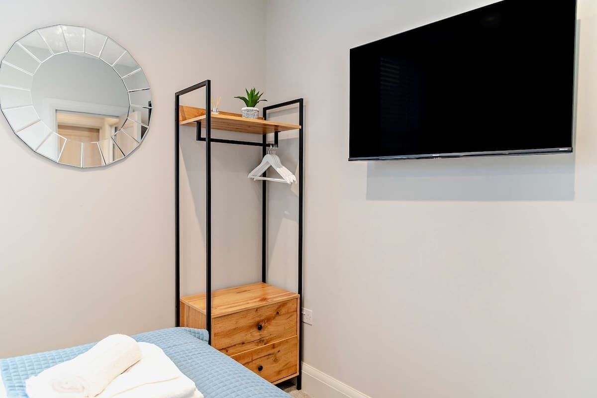Un dormitorio con cama, un espejo redondo, televisión, cómoda de madera y una planta en un estante.