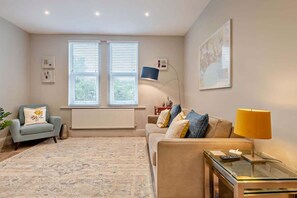 A living room with a sofa, armchair, and a glass coffee table. There are two windows with blinds, a lamp, and a framed picture on the wall.