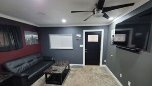A living room with a black leather couch, a coffee table, a ceiling fan, and a flat-screen TV mounted on the wall. - Lake St Clair Boathouse (Harrison Township)