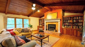 A cozy living room with a fireplace, wooden bookshelves, and a large window. - Murphy Mountain House - Creeks, Firepits & High Speed Internet! (Murphy)
