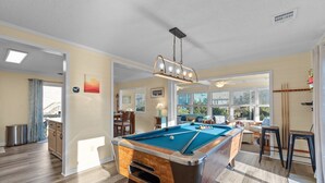A pool table in a well-lit room with wooden flooring, a dining area, and large windows. - 7589- Anchored in the Sand - Avon Beach Cottage | 60 Yards to Beach (Avon)