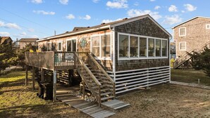 A small, single-story building with a wooden deck and a staircase leading to a covered porch. - 7589- Anchored in the Sand - Avon Beach Cottage | 60 Yards to Beach (Avon)