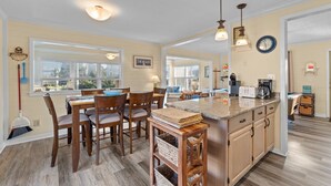 A kitchen with a large island, wooden chairs, and a dining table. There are windows with blinds, a clock, and a sign on the wall. - 7589- Anchored in the Sand - Avon Beach Cottage | 60 Yards to Beach (Avon)