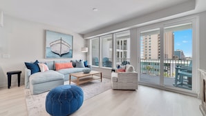 A spacious living room with a large window, a sofa, a coffee table, and a blue ottoman. - "BRAND NEW" BEACHFRONT, OUTDOOR POOL, BEAUTIFUL VIEWS! (Gulf Shores)