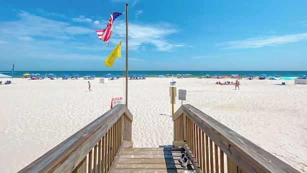 A wooden boardwalk leading to a beach with flags, umbrellas, and beachgoers. - "BRAND NEW" BEACHFRONT, OUTDOOR POOL, BEAUTIFUL VIEWS! (Gulf Shores)