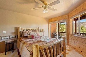 A wooden cabin bedroom with a bed, a ceiling fan, and a window overlooking a mountainous landscape.