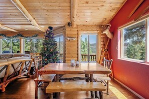 A log cabin interior with a wooden dining table, benches, a Christmas tree, and a deer head mounted on the wall.