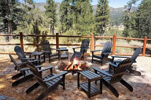 Ein Feuerkorb mit Adirondack-Stühlen darum herum, auf einer Terrasse mit Holzzaun und Bäumen im Hintergrund.