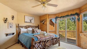A bedroom with a wooden bed, a ceiling fan, a large window with a view, and a wooden deck outside. - The Moose Lodge. Stunning Mtn Views. Spa. Games. (Big Bear Lake)