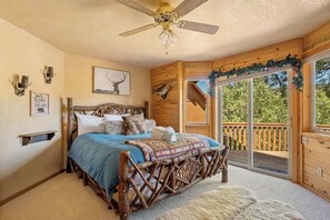 Une chambre avec un lit en bois, un ventilateur de plafond, une grande fenêtre donnant sur un beau paysage et une terrasse en bois à l’extérieur.